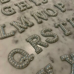 May include: A set of pearl-embellished letters in a silver color. The letters are arranged in alphabetical order, starting with A and ending with Y. The letters are all the same size and are made of a soft, pliable material.
