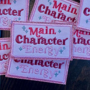May include: Pink embroidered patches with the words "Main Character Energy" in red script. The patches have a light pink background with a glittery pink border and small blue and green diamond accents.