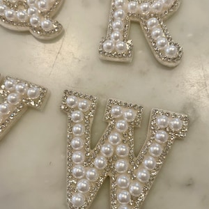 May include: White pearl letters, Q, R, and W, are arranged on a white marble surface. The letters are made of white fabric with white pearls sewn around the edges.