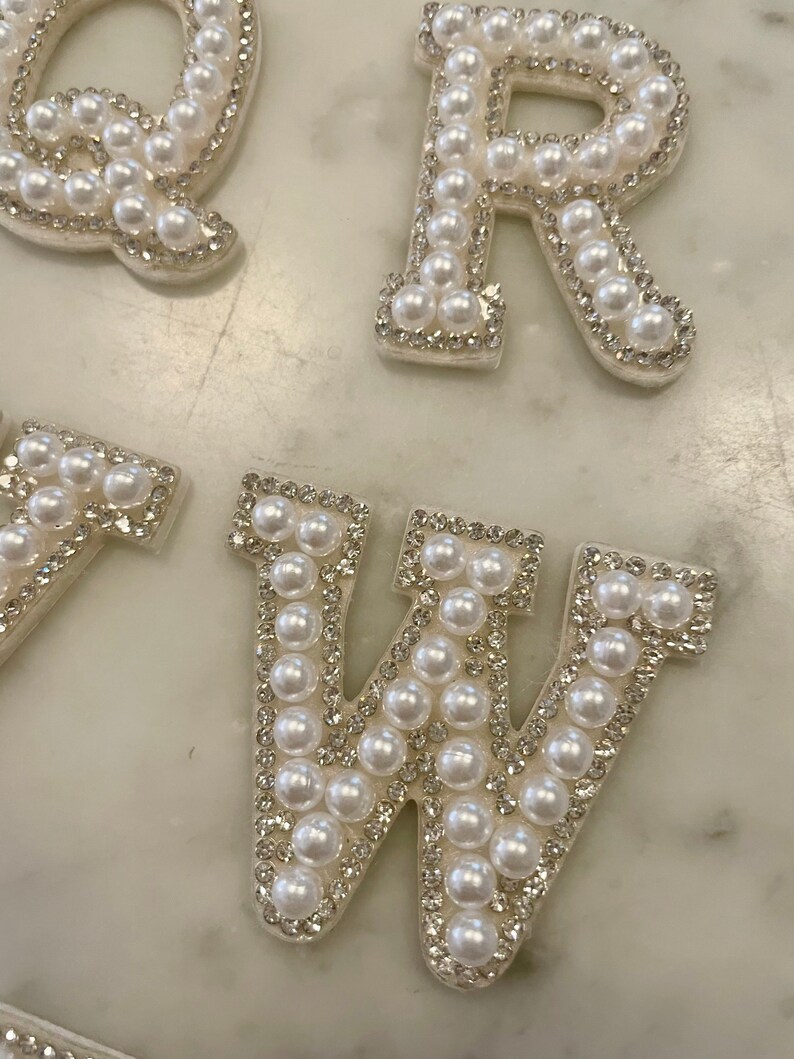 May include: White pearl letters, Q, R, and W, are arranged on a white marble surface. The letters are made of white fabric with white pearls sewn around the edges.