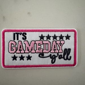 May include: Embroidered patch with the text "IT'S GAMEDAY y'all" in pink and black. The patch has a white background with a pink border and black stars. Perfect for sports fans.