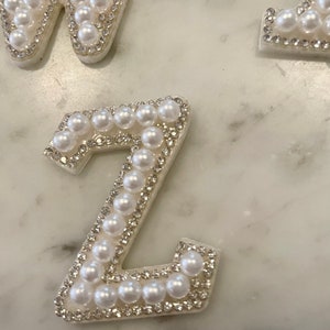 May include: A white letter Z made of pearls and rhinestones. The letter is on a white marble background.