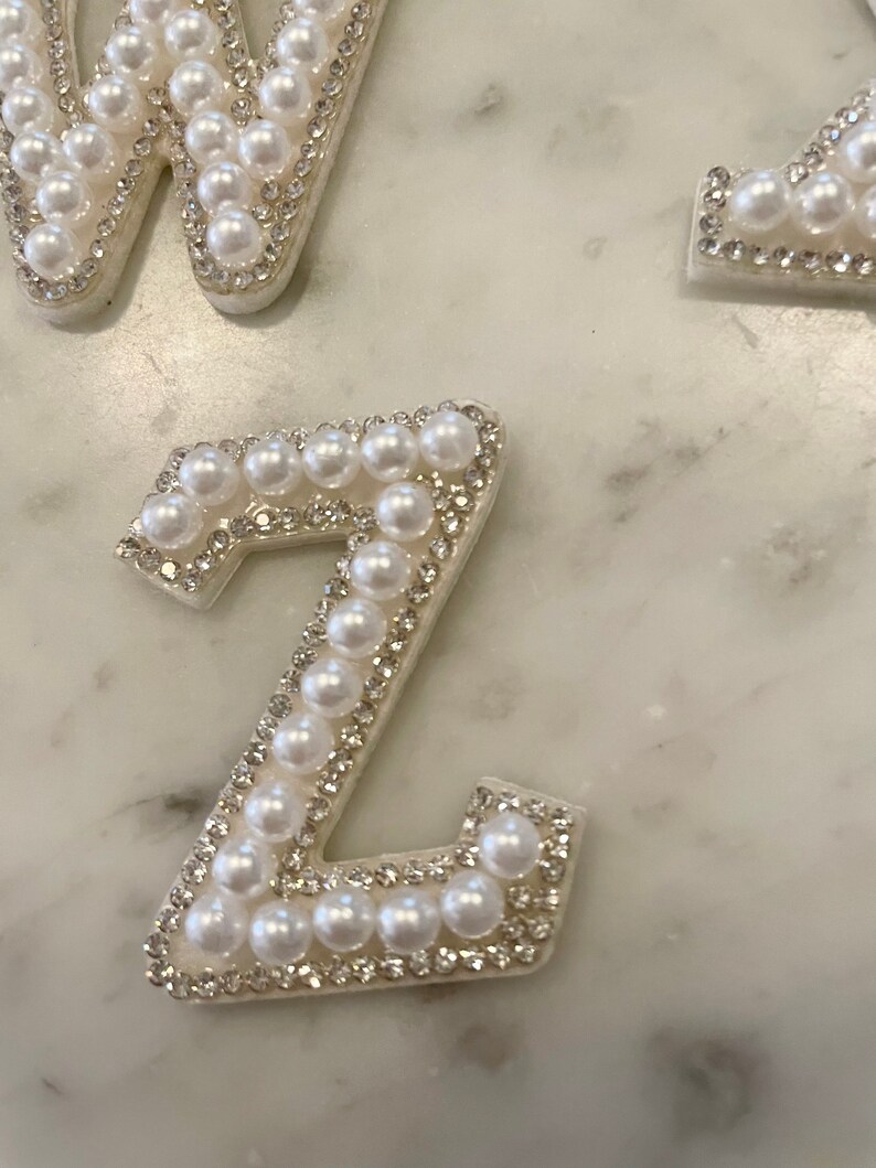 May include: A white letter Z made of pearls and rhinestones. The letter is on a white marble background.