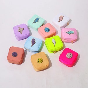 May include: Assortment of small, square zippered pouches in various pastel colors. Each pouch features a different embroidered design, including a smiley face, lightning bolt, and airplane.