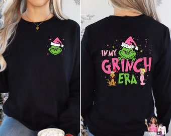 May include: Black sweatshirt featuring a green Grinch face wearing a pink Santa hat on the front. The back of the sweatshirt has the text "IN MY GRINCH ERA" with Grinch and other characters. A festive holiday-themed apparel item.