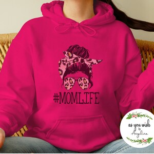 May include: A vibrant fuchsia hoodie featuring a graphic of a woman with a leopard print headband and sunglasses. The text "#MOMLIFE" is printed below the graphic in black. The hoodie has a front pocket and a drawstring hood.