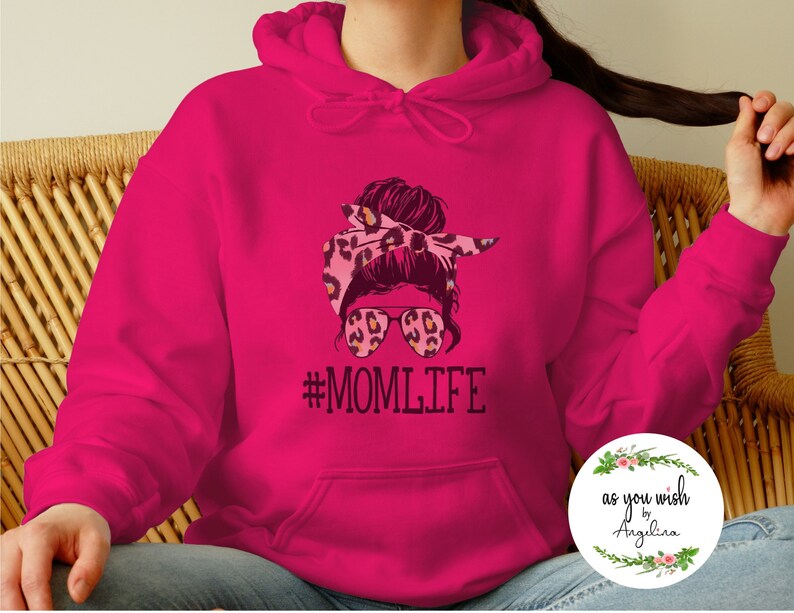 May include: A vibrant fuchsia hoodie featuring a graphic of a woman with a leopard print headband and sunglasses. The text "#MOMLIFE" is printed below the graphic in black. The hoodie has a front pocket and a drawstring hood.