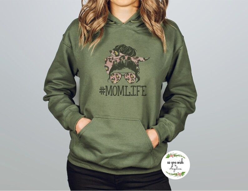 May include: An olive green hoodie with a front pocket and a drawstring hood. The front of the hoodie features a graphic of a messy bun with sunglasses and the text "#MOMLIFE". The graphic includes leopard print accents.