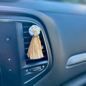 Peut inclure: Un diffuseur de parfum pour voiture en forme de fleur blanche et jaune avec une gland. Il est accroché à une grille d'aération. Le tableau de bord de la voiture est noir.