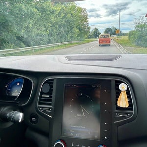 Peut inclure: Int&eacute;rieur d'un tableau de bord de voiture avec un &eacute;cran num&eacute;rique affichant l'heure et la date. Un pompon d&eacute;coratif est suspendu &agrave; la bouche d'a&eacute;ration. Un van vintage orange et blanc est visible sur la route.