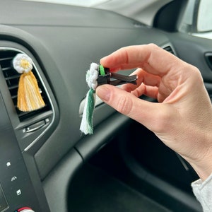 Peut inclure: Diffuseur de voiture &agrave; clipser sur une grille d'a&eacute;ration, avec un pompon blanc, vert et cr&egrave;me. Le clip est noir et est tenu devant le tableau de bord d'une voiture. Un autre diffuseur similaire avec un pompon jaune et blanc est fix&eacute; &agrave; la grille.
