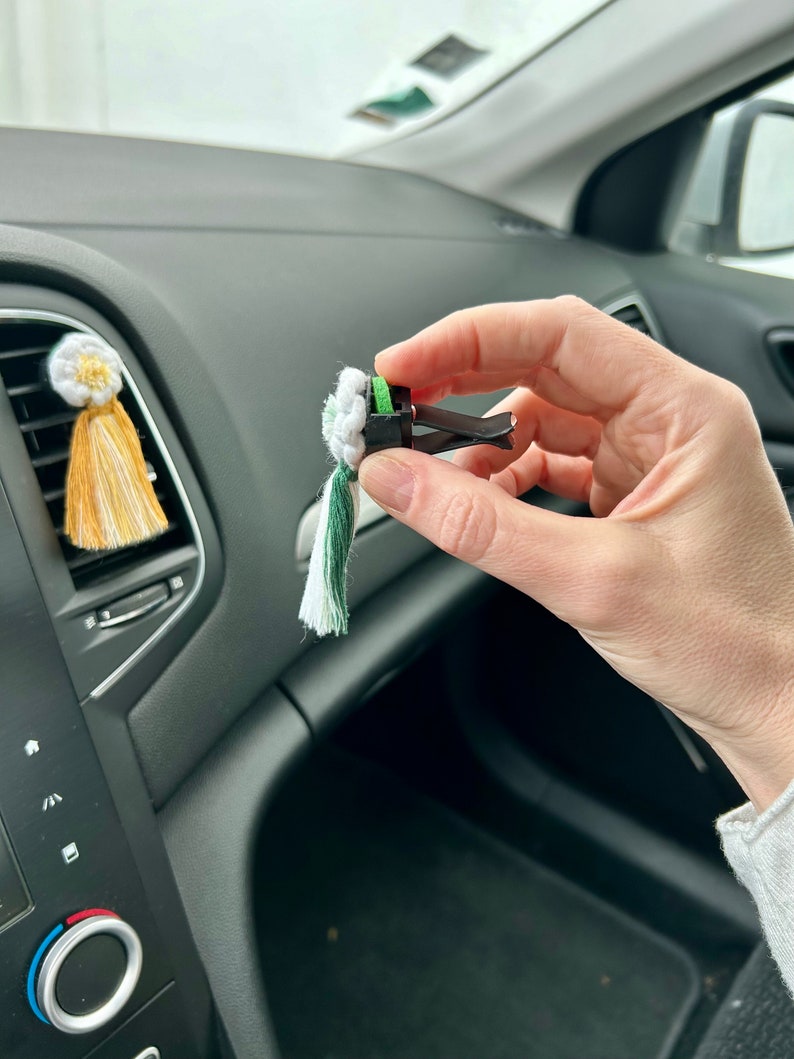 Peut inclure: Diffuseur de voiture &agrave; clipser sur une grille d'a&eacute;ration, avec un pompon blanc, vert et cr&egrave;me. Le clip est noir et est tenu devant le tableau de bord d'une voiture. Un autre diffuseur similaire avec un pompon jaune et blanc est fix&eacute; &agrave; la grille.