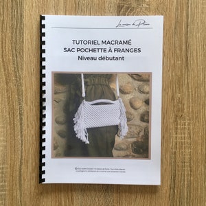 May include: A spiral-bound booklet with the title "TUTORIEL MACRAMÉ SAC POCHETTE À FRANGES Niveau débutant" in French. The cover features a white macrame bag with fringe. The background is a stone wall. The booklet is on a wooden surface.