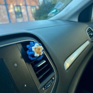 Peut inclure: Un clip de ventilation de voiture en forme de fleur crochetée bleue, blanche et dorée. La fleur est fixée à la grille d'aération de la voiture. Le tableau de bord de la voiture est noir.