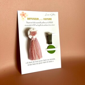Peut inclure: Un diffuseur de voiture avec un design de pompon blanc et rose. Le produit comprend un clip et un disque vert. La carte indique "DIFFUSEUR pour VOITURE" et une citation en fran&ccedil;ais.