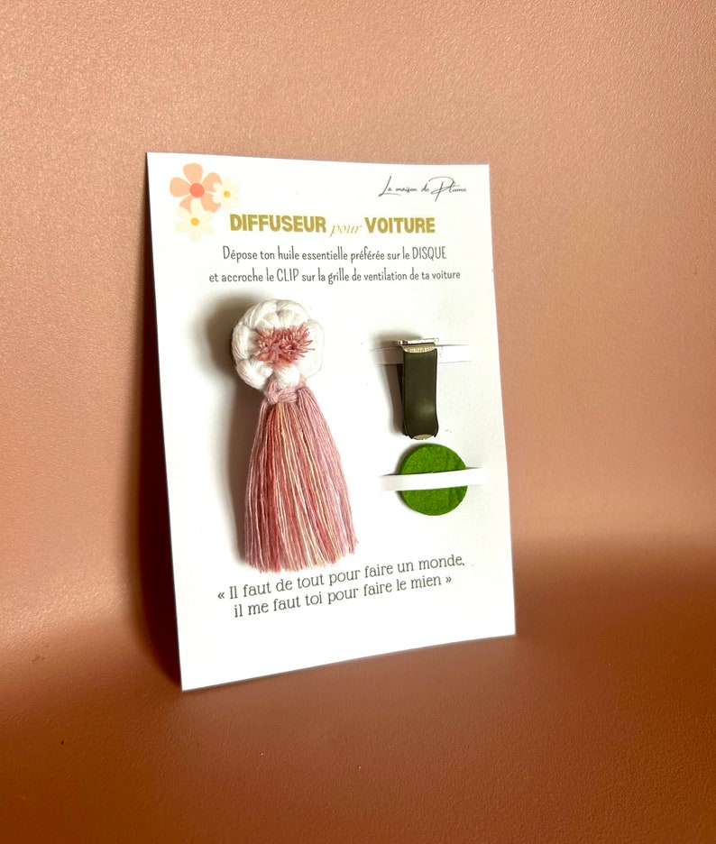 Peut inclure: Un diffuseur de voiture avec un design de pompon blanc et rose. Le produit comprend un clip et un disque vert. La carte indique "DIFFUSEUR pour VOITURE" et une citation en fran&ccedil;ais.