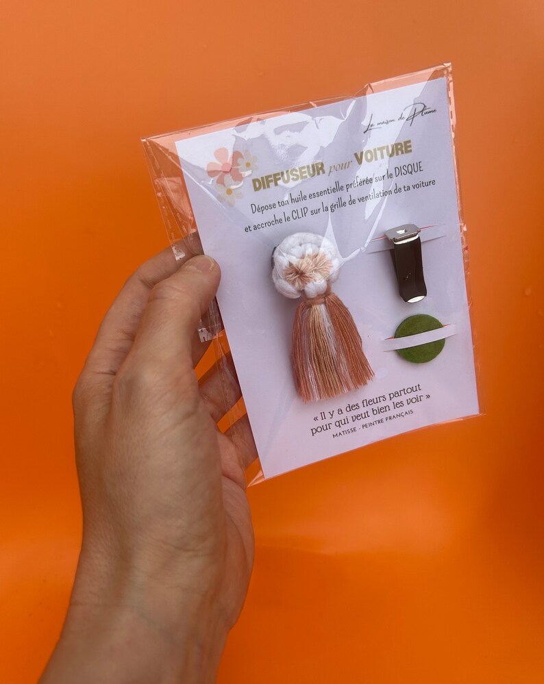 Peut inclure: Un diffuseur de parfum pour voiture pr&eacute;sent&eacute; dans un emballage plastique transparent. Le kit comprend un pompon blanc et beige, un clip noir et un cercle vert. Le texte sur la carte dit "DIFFUSEUR pour VOITURE".