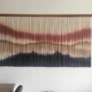Puede incluir: Un gran tapiz de pared con un diseño de paisaje. La obra de arte utiliza hebras verticales de hilo en tonos crema, rojo, marrón y azul oscuro para crear una escena de montaña. Está montado en una barra de madera.