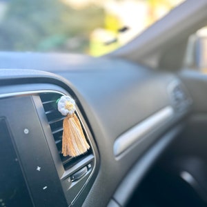 Peut inclure: Un diffuseur de parfum floral blanc et jaune pour voiture, avec une gland en bas. Le tableau de bord de la voiture est noir, et l'arri&egrave;re-plan est flou avec la lumi&egrave;re naturelle.