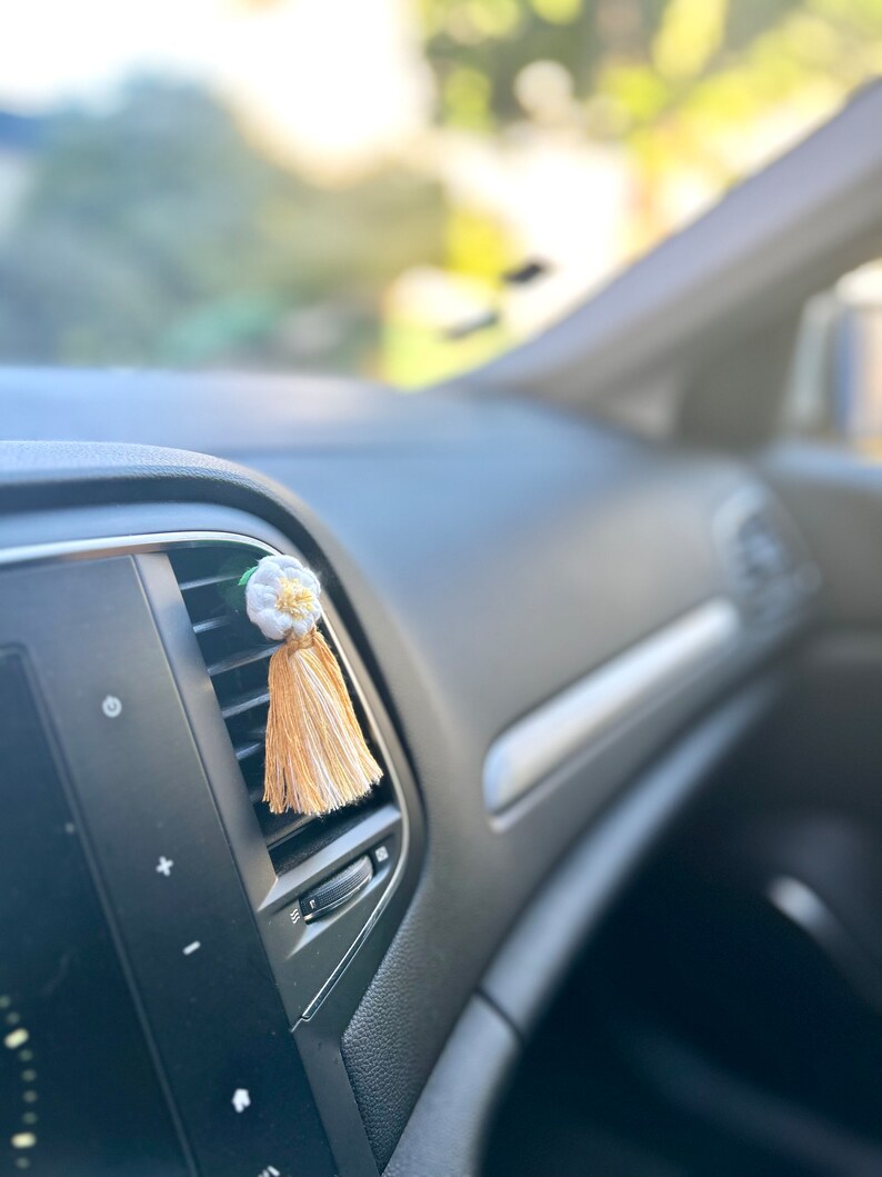 Peut inclure: Un diffuseur de parfum floral blanc et jaune pour voiture, avec une gland en bas. Le tableau de bord de la voiture est noir, et l'arri&egrave;re-plan est flou avec la lumi&egrave;re naturelle.