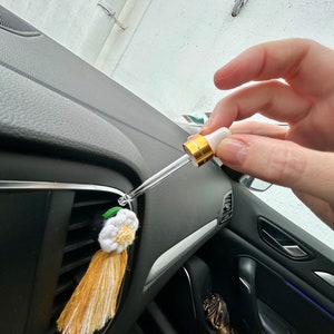 Peut inclure: Diffuseur d'air pour voiture avec un design de pompon et de fleur blanc et jaune. Une main tient une pipette, probablement pour ajouter des huiles essentielles. Le diffuseur est fix&eacute; &agrave; la grille d'a&eacute;ration de la voiture.