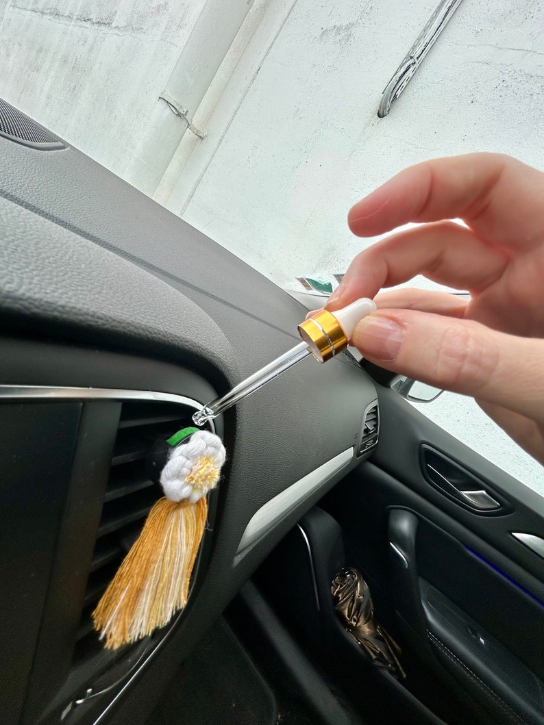 Peut inclure: Diffuseur d'air pour voiture avec un design de pompon et de fleur blanc et jaune. Une main tient une pipette, probablement pour ajouter des huiles essentielles. Le diffuseur est fix&eacute; &agrave; la grille d'a&eacute;ration de la voiture.