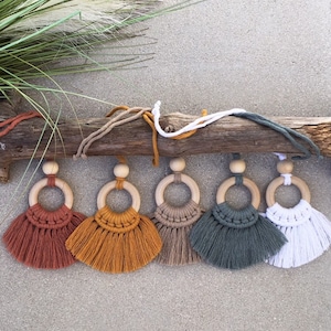 Op de afbeelding: Vijf houten ringen met kleurrijke macramé kwastjes die aan een tak hangen. De kwastjes zijn in de kleuren rood, oranje, bruin, groen en wit.