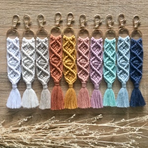 Puede incluir: Una colección de llaveros de macramé hechos a mano en varios colores, incluyendo blanco, gris, melocotón, amarillo, rosa, verde azulado, azul claro y marino. Cada llavero presenta un diseño tejido con una borla y un cierre dorado. Los llaveros están dispuestos sobre una superficie de madera.