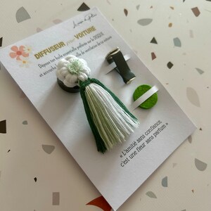 Peut inclure: Diffuseur de voiture avec un pompon blanc et vert et un diffuseur en forme de fleur. Le produit est pr&eacute;sent&eacute; sur une carte avec du texte, dont "DIFFUSEUR POUR VOITURE".