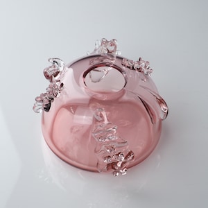 May include: A pink glass bowl with clear glass accents. The bowl has a round shape and a wide opening. The accents are in the shape of small, irregular swirls.
