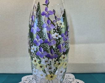 Puede incluir: Un jarrón de vidrio transparente con diseños florales pintados a mano. El jarrón presenta flores moradas, blancas y amarillas con hojas verdes. El jarrón está colocado sobre un tapete de ganchillo blanco.