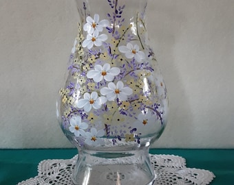 Puede incluir: Un florero de vidrio transparente con borde acampanado y cuerpo redondeado, pintado a mano con un diseño floral que presenta flores blancas de cinco pétalos, pequeñas flores amarillas y delicadas ramas moradas sobre la superficie.