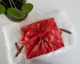 Könnte beinhalten: Ein rotes Stoffgeschenkpapier mit einem goldenen Glitzermuster aus Weihnachtsbäumen. Der Stoff ist mit einer Schleife gebunden und liegt auf einer weißen, flauschigen Oberfläche.