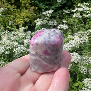 May include: A polished, tumbled gemstone featuring a smooth, rounded surface with a mottled appearance. The stone displays a blend of deep magenta and pink inclusions set against a translucent, cloudy grey and white mineral matrix throughout.