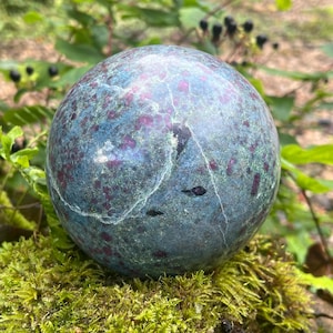 May include: A spherical gemstone with a polished surface, displaying a mottled pattern. The stone's colours include teal, burgundy, and green. It rests on green moss, with a blurred natural backdrop. The sphere is approximately 8 cm in diameter.