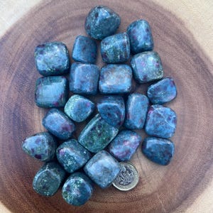 May include: A collection of tumbled stones in shades of blue and green, with flecks of red. The stones are smooth and rounded, and a coin is included for size reference. The stones are arranged on a wooden surface.