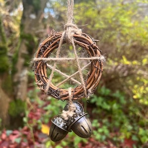 May include: A rustic, circular wreath crafted from woven brown twigs, featuring a five-pointed star design made with twine. Two bronze-toned acorn-shaped bells are suspended below. The wreath hangs from a natural twine cord, ideal for display.