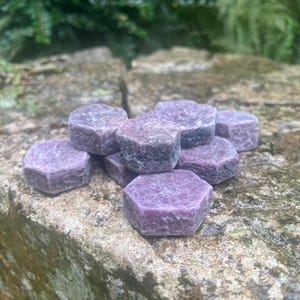 May include: A collection of small, raw, hexagonal-shaped ruby crystals with a deep magenta and purple hue. The stones feature a natural, unpolished texture with visible crystalline structures and varying shades of dark red and violet throughout.