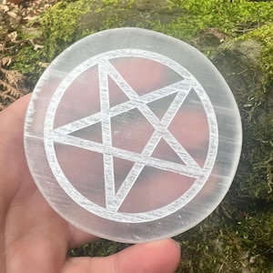 May include: A circular, clear selenite crystal plate featuring a white pentagram design etched on its surface. The crystal is held in a hand, with a mossy green background.