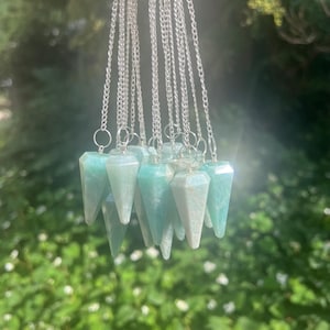 May include: A collection of multiple light blue-green gemstone pendulums, each with a faceted conical shape and a silver-toned metal jump ring attached to a silver-toned chain. The stones exhibit natural variations in colour and opacity.