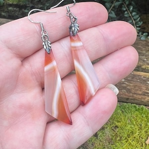 May include: Dangle earrings with triangular agate stones in orange, white, and clear. The earrings have silver-coloured hooks and leaf accents at the top. The design creates a natural and elegant look, suitable for everyday wear.