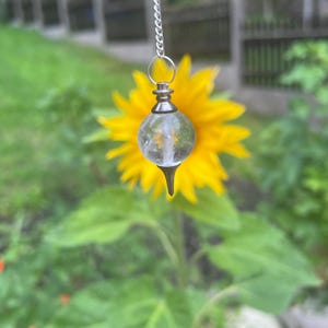 May include: A clear spherical crystal pendulum featuring a silver-toned metal cap and a pointed base, suspended from a delicate silver-coloured metal chain. The crystal contains visible internal inclusions and is centred in the frame.