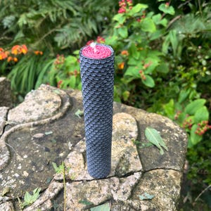 May include: A black and grey honeycomb beeswax candle with a red centre. The candle is sitting on a stone surface.