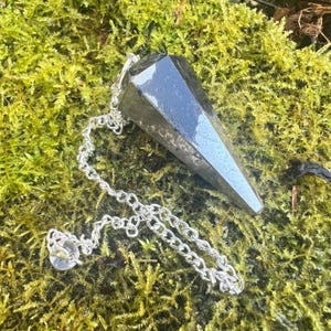 May include: A dark, faceted pendulum with a silver chain and a clear crystal at the end. The pendulum is resting on a bed of green moss, creating a natural and earthy aesthetic. The geometric shape of the pendulum contrasts with the organic background.