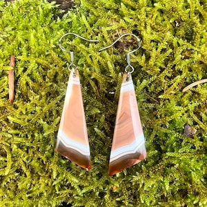 May include: A pair of earrings with a triangular design. The earrings display a layered pattern of brown, white, and peach bands. They hang from silver-coloured hooks. The backdrop is a bed of green moss.