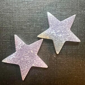 May include: Two sparkly, iridescent purple star-shaped ornaments. The stars are made of a translucent material with a glittery finish.