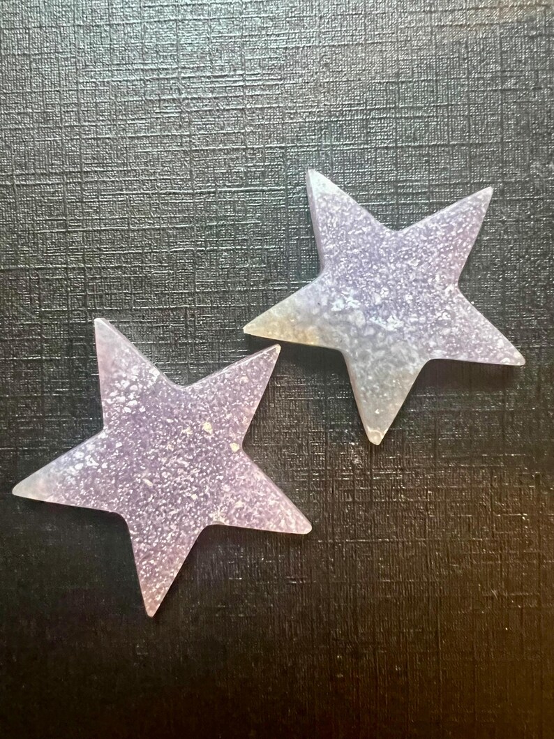 May include: Two sparkly, iridescent purple star-shaped ornaments. The stars are made of a translucent material with a glittery finish.