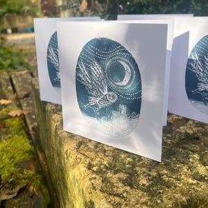 May include: A set of white greeting cards featuring a blue and white illustration of an owl in flight against a night sky with a crescent moon and a church. The cards are displayed on a mossy stone surface.