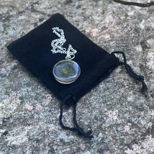 May include: A circular pendant with a dark, mottled green and black interior encased in a clear, smooth dome. The pendant is attached to a silver-toned metal chain and rests on a small, black fabric drawstring pouch with black cord ties.
