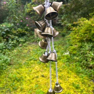May include: A string of brass bells hanging from a white cord. The bells are arranged in a cascading pattern, with the largest bell at the top and the smallest bell at the bottom. The bells are all in focus, and the background is blurred.
